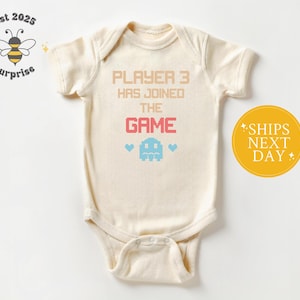 May include: Cream-colored baby onesie with the text "PLAYER 3 HAS JOINED THE GAME" in peach and pink. A pixelated ghost and hearts are below the text. The onesie has short sleeves and a snap closure.