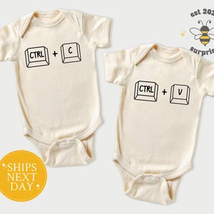 May include: Two cream-colored baby onesies. One features the text "CTRL + C" and the other "CTRL + V" in black. A yellow circle with the text "SHIPS NEXT DAY" is visible. The image also includes a bee graphic with the text "est 2025 surprise".