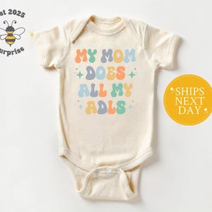 May include: Cream-colored baby onesie with the text "MY MOM DOES ALL MY ADLS" in colorful, retro-style lettering. The onesie has short sleeves and a snap closure. A yellow circle with the text "SHIPS NEXT DAY" is on the right.