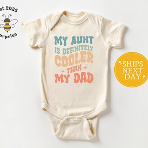 May include: Cream-colored baby onesie with the text "MY AUNT IS DEFINITELY COOLER THAN MY DAD" in retro-style lettering. The onesie has a snap closure and short sleeves. A yellow circle with the text "SHIPS NEXT DAY" is on the right.