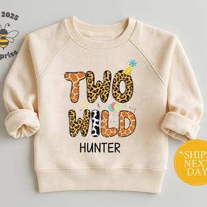May include: Cream-colored sweatshirt with the text "TWO WILD HUNTER" in a mix of animal print and giraffe patterns. A party hat and balloons are included in the design. The sweatshirt has long sleeves and a round neckline.