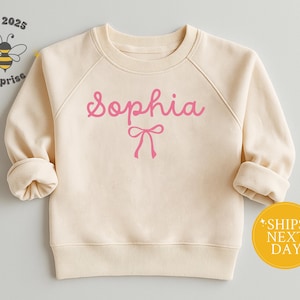 May include: Cream-colored children's sweatshirt with the name "Sophia" in pink script and a pink bow graphic. The sweatshirt has long sleeves with rolled cuffs. A yellow circle with text is visible.