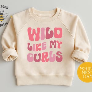 May include: Cream-colored sweatshirt with the text "WILD LIKE MY CURLS" in shades of pink. The sweatshirt has long sleeves with rolled cuffs. A yellow circle with the text "SHIPS NEXT DAY" is on the bottom right.