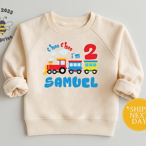May include: Cream-colored sweatshirt with a colorful train graphic. The design includes a red train, blue and yellow train cars, the number 2, and the name "Samuel" in blue. The phrase "Choo Choo I'm 2" is also included.
