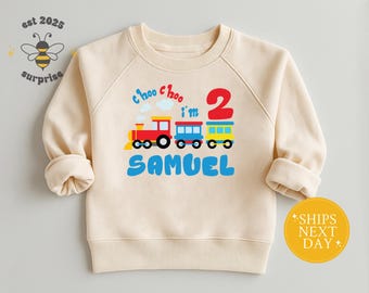 May include: Cream-colored sweatshirt with a colorful train graphic. The design includes a red train, blue and yellow train cars, the number 2, and the name "Samuel" in blue. The phrase "Choo Choo I'm 2" is also included.