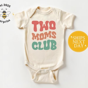 May include: Cream-colored baby onesie with the text "TWO MOMS CLUB" in retro-style lettering. The text is in shades of red, peach, and teal. A yellow circle with text is on the right. A bee logo is on the left.