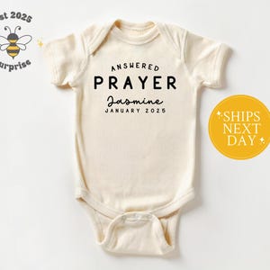 May include: Cream-colored baby onesie with the words "ANSWERED PRAYER" in black, followed by "Jasmine" and "JANUARY 2025". A yellow circle with "SHIPS NEXT DAY" is on the right. The top left corner has a bee logo.