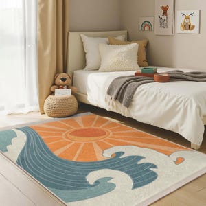 Puede incluir: Una alfombra azul y naranja con un diseño de sol y olas. La alfombra está sobre un suelo de madera clara delante de una cama blanca con una manta de lana gris.