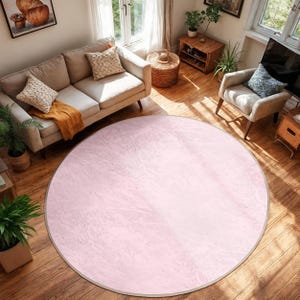 Può includere: Un grande tappeto rotondo con un motivo rosa chiaro simile al marmo. Il tappeto ha un sottile bordo grigio ed è posizionato su un pavimento in legno. Il tappeto è un elemento decorativo in un soggiorno, con un divano, una poltrona e altri mobili visibili.