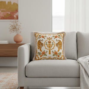 May include: A square throw pillow with a cream-colored background featuring a symmetrical pattern of two tigers, orange floral motifs, and a central gold crest with a crown. The pillow is finished with a light beige fringe along all four edges.