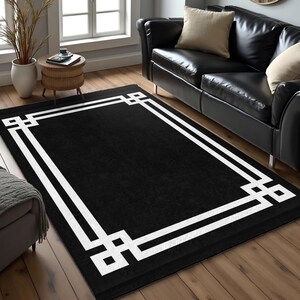 Puede incluir: Una alfombra rectangular negra con un borde geométrico blanco. La alfombra está colocada sobre un suelo de madera en un salón, con un sofá de cuero negro visible al fondo.