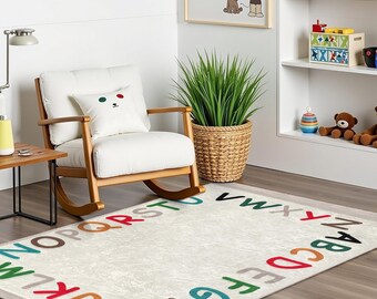 May include: A white rocking chair with a white cushion and a green and red bear pillow sits on a cream colored rug with colorful letters of the alphabet printed on it.