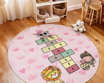 May include: A pink round play mat with a hopscotch game featuring an owl, a lion, and a girl. The mat has a pink background with white crowns, hearts, and wings. The hopscotch game has numbers 1 through 11. The words "love" are repeated throughout the mat.