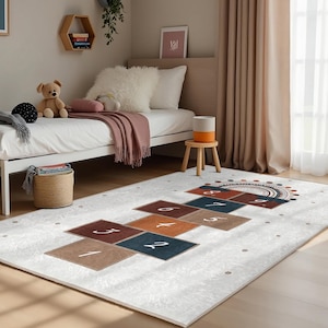 Può includere: Un tappeto da gioco bianco e grigio con un design di hop scotch con i numeri da 1 a 9. Il tappeto ha uno schema di colori marrone, arancione e blu. Il tappeto si trova in una stanza con un letto bianco e uno sgabello di legno.