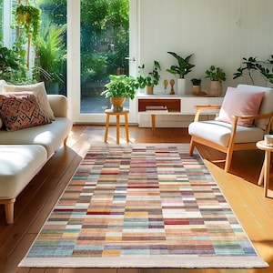 Op de afbeelding: Een kleurrijk rechthoekig vloerkleed met een geometrisch patroon van meerkleurige strepen. Het vloerkleed ligt op een houten vloer in een woonkamer met meubels en planten.