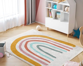 May include: A white and grey rainbow rug with yellow, pink, red, and blue stripes. The rug is on a wooden floor in a room with a white bookshelf and a pink wall.