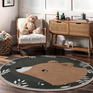 May include: A round area rug featuring a brown bear face on a dark green background, surrounded by white leaf accents. The rug is placed on a wooden floor in a room with a teddy bear, a rocking chair, and a wooden cabinet.