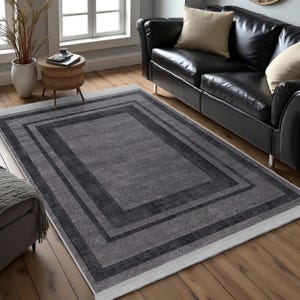 May include: A gray area rug with a black border. The rug has a subtle pattern and a white fringe around the edges.