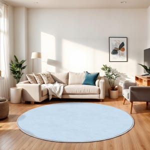 Könnte beinhalten: Ein hellblaues, rundes Teppich mit weißem Rand liegt auf einem Holzboden. Der Teppich ist der Mittelpunkt eines Wohnzimmers mit beigem Sofa, Pflanzen und Kunstwerken im Hintergrund. Der Teppich hat einen Durchmesser von ca. 200 cm.