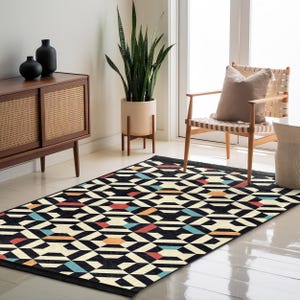 May include: Geometric patterned area rug with a black border. The rug features a repeating pattern of black, cream, red, orange, and blue geometric shapes. The rug is placed on a light-colored floor in a well-lit room.
