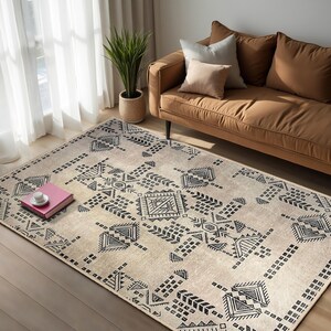 May include: A beige area rug with a black geometric pattern. The rug features a repeating design of squares, triangles, and lines. The rug is laid out on a wooden floor in a living room setting.