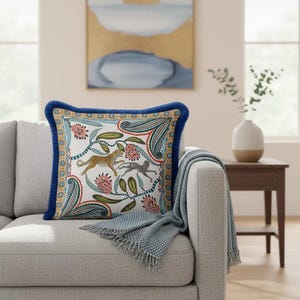 May include: A square throw pillow featuring a white background with a colorful pattern of a leopard, a monkey, and floral elements. The pillow is framed by a decorative border and finished with a dark blue velvet-like piping along the edges.