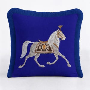 Op de afbeelding: Een vierkant koningsblauw sierkussen met een centraal borduurwerk van een wit paard in beweging. Het paard draagt een bruin en goudkleurig zadel en hoofdstel met een klein gouden embleem. Het kussen is afgewerkt met een bijpassende blauwe franje.