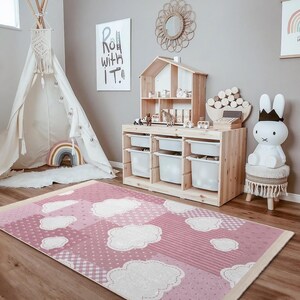 May include: A pink and white cloud-themed rug with a checkered border. The rug is in a playroom with a teepee, a wooden toy shelf, and a white bunny toy.