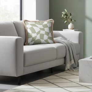 May include: A square throw pillow featuring a sage green and white wavy checkerboard pattern. The pillow is finished with a textured, light brown fringe trim along all four edges, providing a distinct border to the soft, patterned fabric surface.