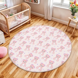 Puede incluir: Una alfombra redonda de color crema con un patrón repetido de osos de peluche rosas y corazones morados. La alfombra está colocada sobre un suelo de madera, con una cuna visible en el fondo. Una alfombra suave y decorativa para una guardería.