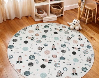 Puede incluir: Una alfombra redonda de color blanco roto con un diseño espacial. El diseño incluye cohetes, planetas, estrellas y constelaciones en tonos azules, rosas y negros. La alfombra está colocada sobre un suelo de madera. Diámetro: aprox. 150 cm.