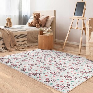 Può includere: Un tappeto bianco e rosa con un motivo ripetuto di scimmie rosa e palle blu e bianche. Il tappeto è su un pavimento di legno in una stanza con un letto e un tavolino di legno.
