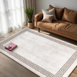 Könnte beinhalten: Ein weißer Teppich mit einem braunen griechischen Schlüsselmuster als Bordüre. Der Teppich liegt auf einem Holzboden und im Hintergrund befindet sich ein braunes Sofa.