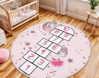May include: A round pink rug with a hopscotch design, featuring illustrations of ballerinas and stars. The rug has a hopscotch grid with numbers 1 through 10. The rug is designed for a child's room.