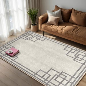 May include: A white area rug with a geometric pattern in a dark grey colour. The rug has a subtle texture and a border of the same geometric pattern. The rug is laid out on a wooden floor in a living room setting.