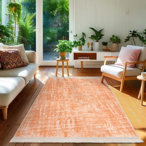 Puede incluir: Una alfombra de área de color naranja claro con un aspecto desgastado y texturado. La alfombra tiene un borde blanco y está colocada sobre un suelo de madera en un salón.