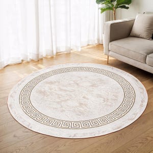 Op de afbeelding: Een rond tapijt met een crèmekleurige achtergrond en een decoratieve rand. De rand heeft een zich herhalend geometrisch patroon in donkerbruin. Het tapijt ligt op een houten vloer en op de achtergrond is een bank te zien.