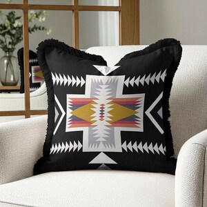 May include: A square throw pillow with a black base and a central geometric pattern featuring white, yellow, pink, and grey triangles and diamonds. The pillow is finished with a black fringed edge along all four sides, creating a textured border.