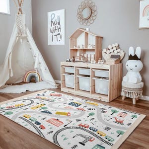 May include: A white and gray play rug with a colorful road and train track design. The rug features cars, trucks, trains, and buildings. The rug is in a playroom with a teepee, a toy shelf, and a white bunny rabbit.