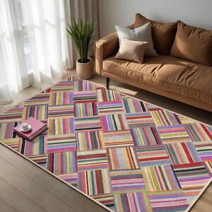 Op de afbeelding: Een kleurrijk gestreept vloerkleed met een geometrisch patroon in tinten roze, paars, groen, blauw, geel, oranje en bruin. Het vloerkleed ligt op een houten vloer in een woonkamer.