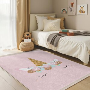Può includere: Un tappeto a forma di unicorno rosa e oro con un corno glitterato e dettagli floreali. Il tappeto è su un pavimento di legno in una camera da letto con un letto bianco e un peluche.