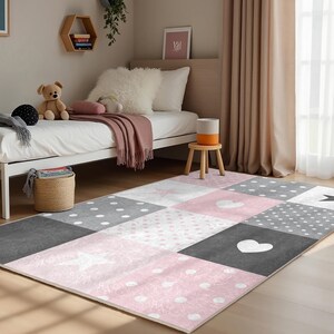 May include: A pink and gray patchwork rug with white polka dots, stars, and hearts. The rug is in a child's bedroom with a white bed and a wooden stool.