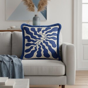 May include: A square throw pillow featuring a cream-colored base with a bold, dark blue abstract wavy pattern radiating from the center. The pillow is finished with a dark blue fringe trim along all four edges, providing a textured border.