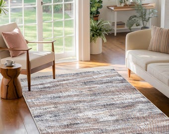 Puede incluir: Una alfombra de área de color marrón claro y gris con un borde blanco. La alfombra tiene un patrón abstracto desgastado. La alfombra está en un ambiente de sala de estar con un sofá blanco y una silla de madera.