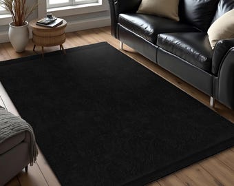 May include: A black area rug with a subtle texture. The rug is laid out on a hardwood floor in a living room setting with a black leather sofa in the background.