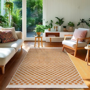 May include: A checkered area rug with a white background and orange squares. The rug is in a living room with a white couch, a white armchair, and plants.