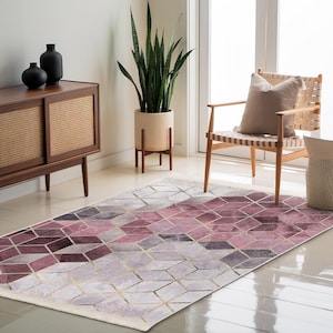 May include: A geometric patterned rug with a cream background and pink and gray diamond shapes. The rug has gold accents and is laid out on a hardwood floor in a living room setting.