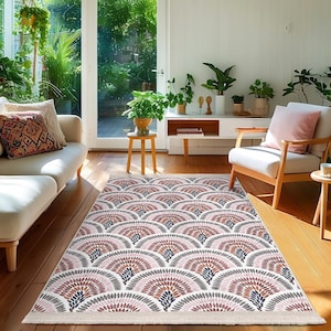 Puede incluir: Una alfombra rectangular con un patrón repetitivo en forma de abanico en tonos rosa, marrón, gris y azul marino. La alfombra está colocada sobre un suelo de madera en un salón, con una franja blanca en los bordes.