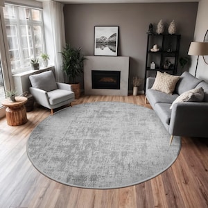 May include: A large, round, light gray rug with a distressed pattern dominates the room's hardwood floor. The rug's design features a mottled texture, creating a soft, inviting look. The rug is a central element in a living room setting.