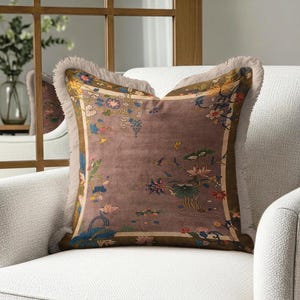 May include: A square throw pillow featuring a muted mauve background with a decorative border and a central floral pattern in shades of blue, yellow, and green. The pillow is finished with a light beige fringe trim along all four edges.